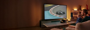 Un père et son fils souriant en regardant une course automobile à grande vitesse à la télévision, avec l’éclairage connecté WiZ derrière l’écran et sur les étagères à proximité.