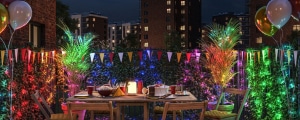 Outdoor table set for a party at night with colorful lights, balloons, bunting, and food, surrounded by greenery and apartment buildings in the background.