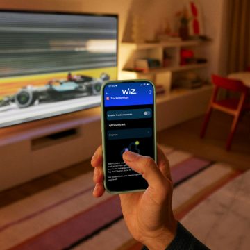A hand holding a phone using the WiZ App. A TV showing a race lit by WiZ Smart Lighting in the background.