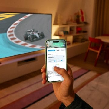 Une main tenant un téléphone et utilisant l'application WiZ. Un téléviseur montrant une course avec l'éclairage connecté WiZ en arrière-plan.