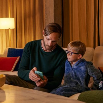 Een vader laat zijn telefoon zien aan zijn zoontje op de bank. Een lamp met een slimme WiZ lamp en warm licht.